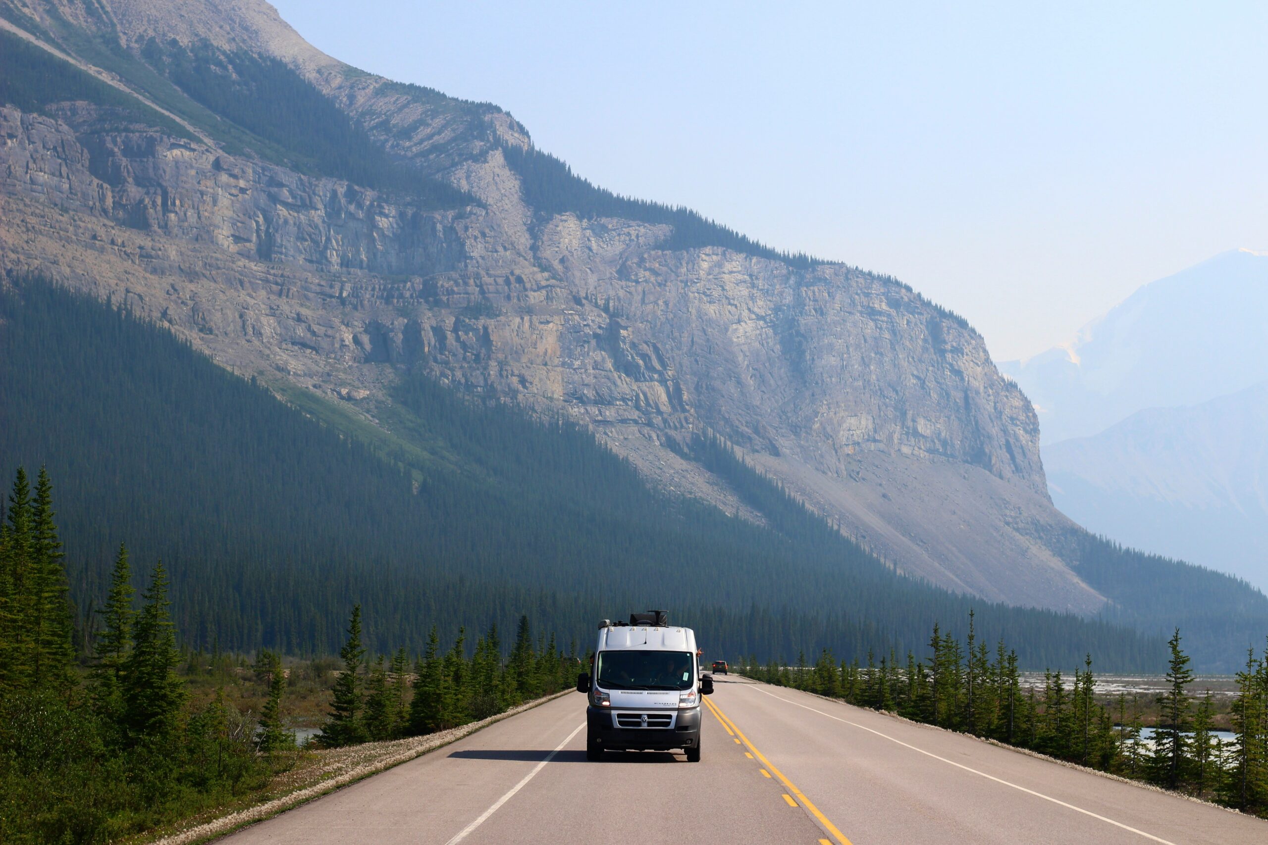 Motor home driving on an empty road with a mountain and trees in the background.